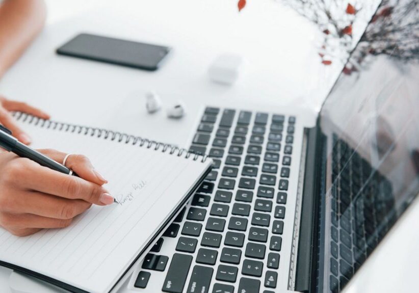 Using laptop and notepad. Young female freelancer working indoors in the office at daytime.